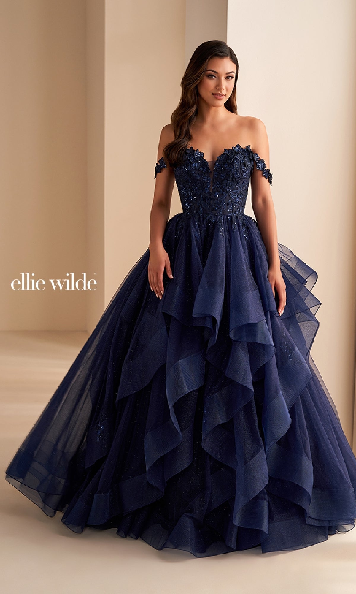 A woman wears the Ellie Wilde Long Designer Prom Ball Gown EW35084—an off-the-shoulder, navy blue gown with floral embellishments and layered glitter tulle—standing indoors against a neutral background.